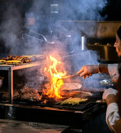 A chefs hands are grilling food with flames rising in a busy restaurant kitchen setting.